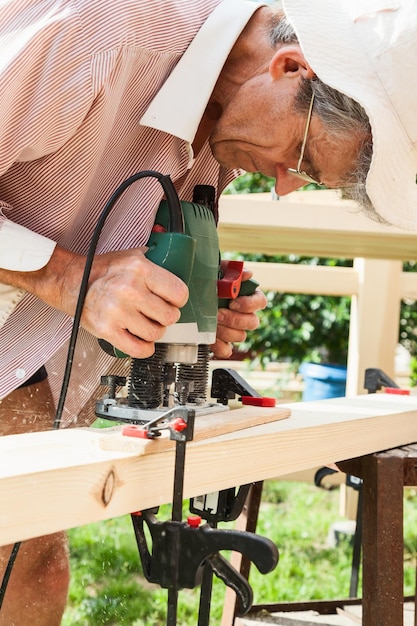 Photo an elderly man at his summer cottage in a panama hat works with a green manual milling machine over lumber against a background of bushes