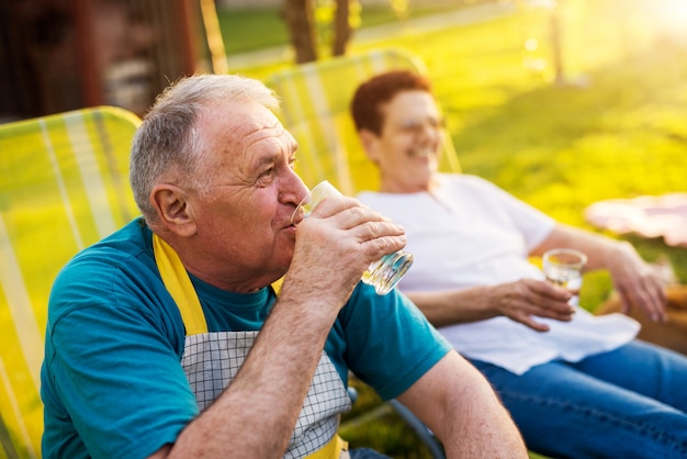 Elderly man is drinking water and looking into the distance while his woman is sitting beside him.