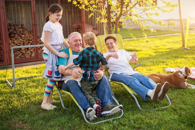 Elderly man is holding his little grandson while his granddaughter is standing behind him and his wife sitting beside him on a beautiful day.