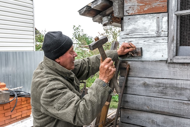 An elderly man is repairing an old wooden shed He nails the board to the wall