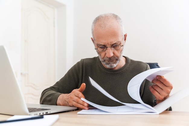 Elderly man leafing through the pages of a notebook and working on a laptop