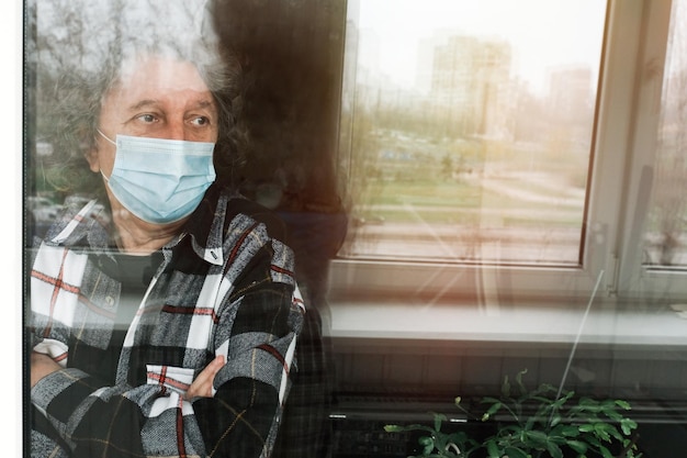 An elderly man in a protective mask on selfisolation at home looks out the window Quarantine due to the global coronavirus pandemic Lifestyle