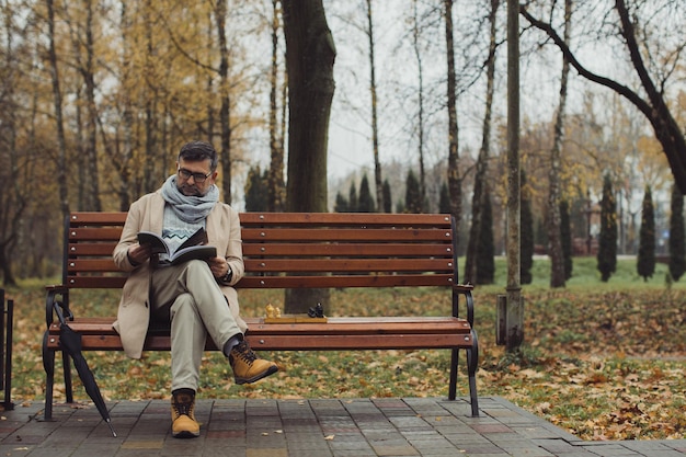 An elderly man reads a magazine while sitting on a bench in an autumn park A man is waiting for his friend to play chess