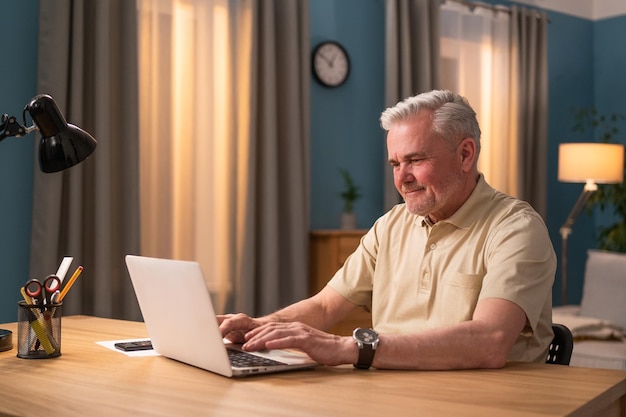 Elderly man sitting at desk with laptop in the evening grandfather talks to grandchildren online