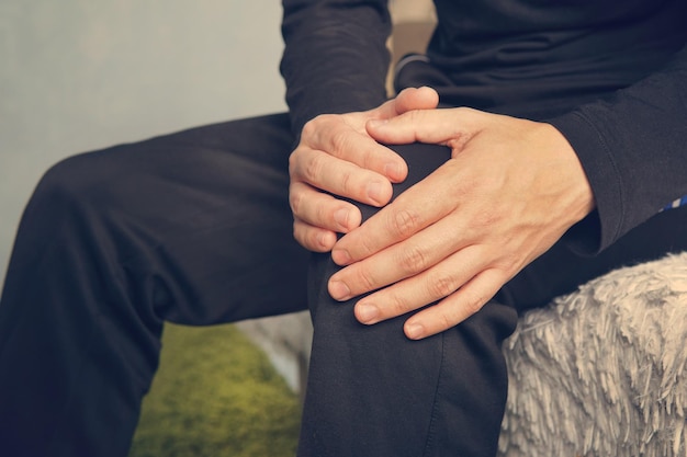 Elderly man sitting on a sofa in the living room at home and touching his knee by the pain