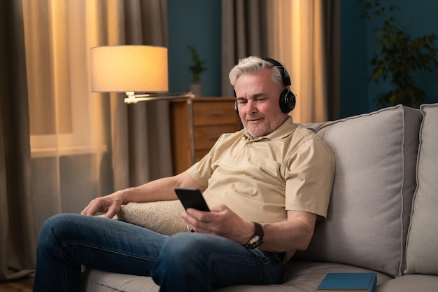 An elderly man sitting on a sofa in the living room listening to music from his phone on headphones
