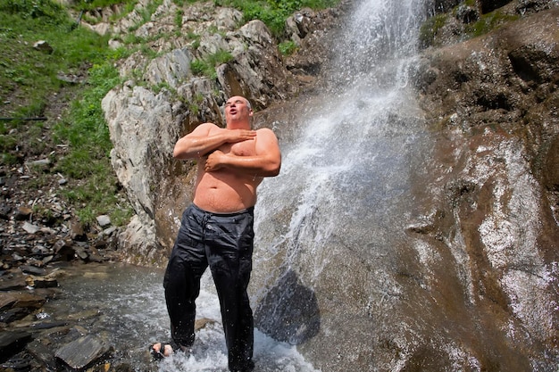 An elderly man stands at a mountain waterfall enjoys the moment