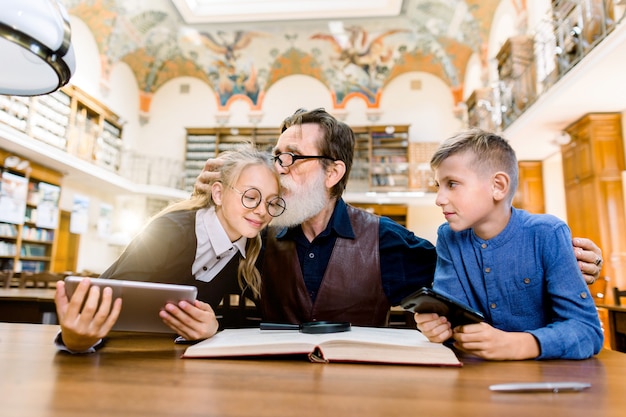 Elderly man teacher and his teen students, boy and girl, sitting at the table in library