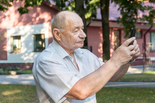 An elderly man walks alone in the park in the summer A modern pensioner businessman in a white shirt and trousers takes pictures with a camera in a mobile phone