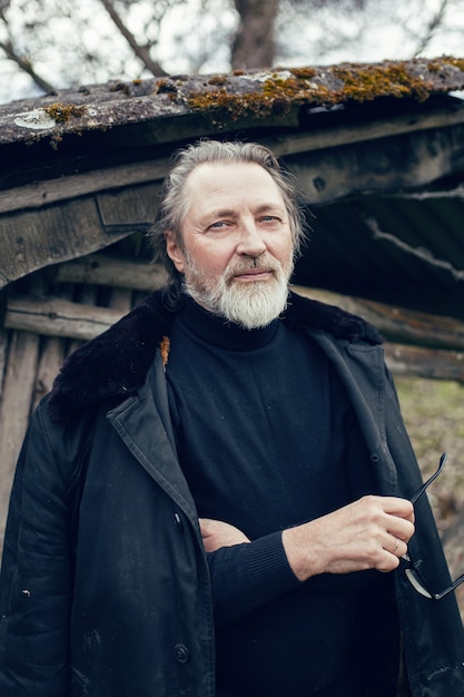 Elderly man with a beard stands in the village near a wooden shed in a sheepskin coat and glasses