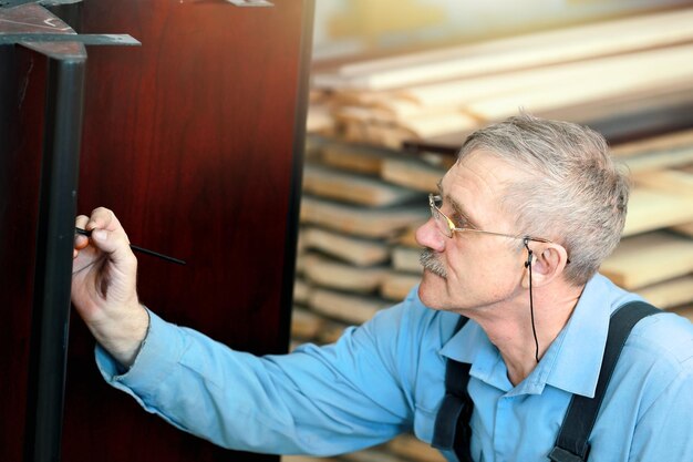 An elderly man with glasses and gray hair works with wood in a carpentry shop