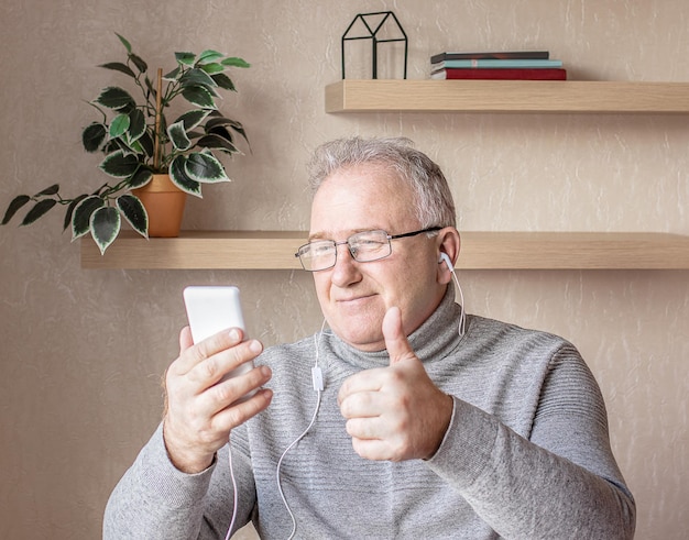Пожилой мужчина в очках, наушниках и смартфоне в руке изучает онлайн-курсы с помощью компьютера Концепция дистанционного обучения