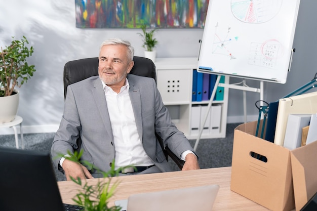 An elderly man with gray hair sits on a chair at an office desk\
next to him a box of packed things