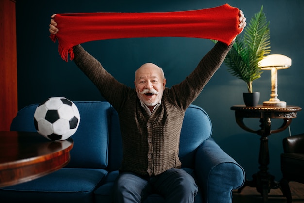 Elderly man with red scarf and ball watching TV, football fan. Bearded mature senior poses in living room, old age people leisures