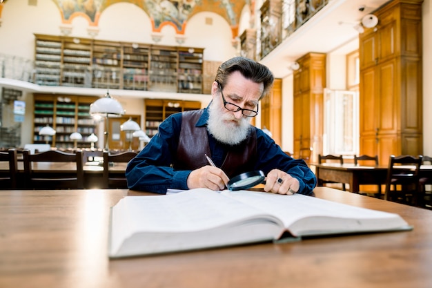 Uomo anziano con barba bianca e occhiali lavorando in un'antica biblioteca con libri, seduto al tavolo d'epoca. istruzione, concetto di biblioteca