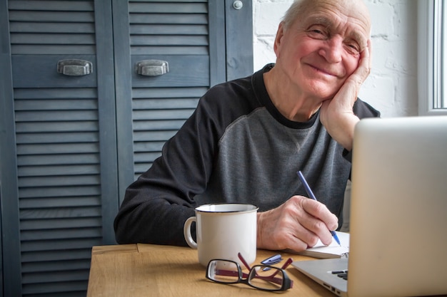 Elderly man working on laptop, smiling, looking at screen, drinking coffee.