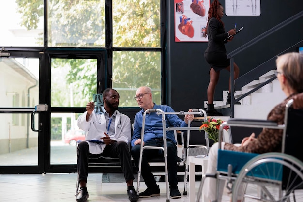 Elderly patient with walking frame sitting in hospital lobby analyzing radiography with physician doctor discussing disease diagnosis during consultation. Patients waiting in reception area