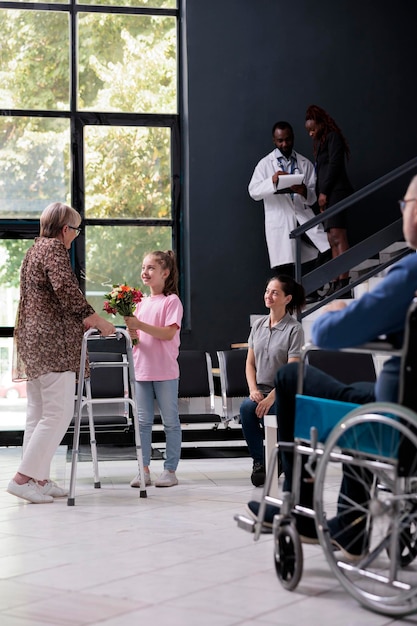 Foto paziente anziano con deambulatore in piedi nella reception dell'ospedale durante la consultazione della visita di controllo. la nipote accoglie la nonna con un mazzo di fiori. servizio e concetto di medicina