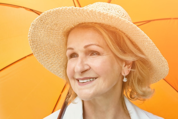 Elderly pretty woman about 60 years old in hat is smiling while standing under orange umbrella outdoor during day.