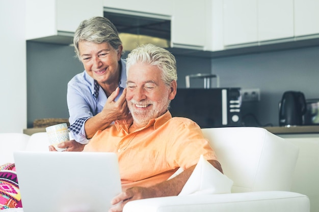 Elderly real lifestyle people using computer at home Leisure indoor activity with man and woman enjoying laptop and internet connection sitting on the sofa with kitchen in background