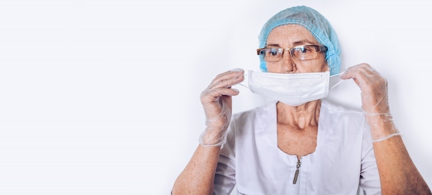 Elderly sad tired mature woman doctor or nurse in a white medical coat, gloves, puts on face mask wearing personal protective equipment isolated. Healthcare and medicine concept. Covid-19 pandemic