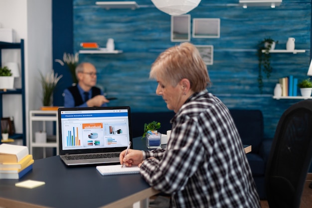 Elderly senior woman taking notes in notebook while working on laptop