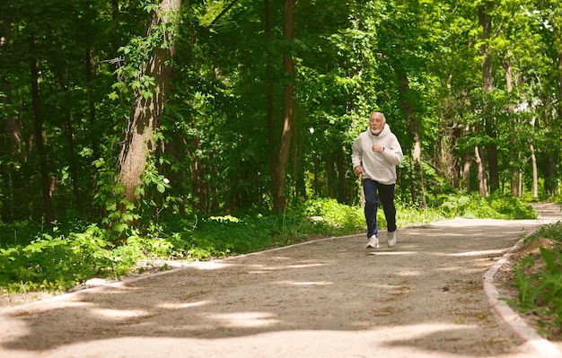 Elderly sporty man running in green forest during morning workout, copy space. Healthy and active lifestyle at any age concept