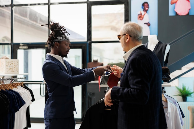 Elderly stylish client discussing tie fabric with store worker, looking at new fashion collection in modern boutique. Senior customer shopping for casual wear, buying fashionable clothes.