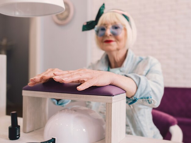 elderly stylish woman in blue sunglasses and denim jacket sitting at manicure salon studio ap