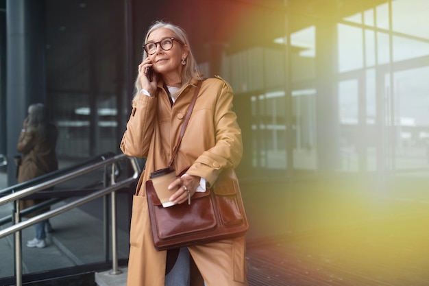 Пожилая стильная женщина разговаривает по мобильному телефону рядом с торговым центром