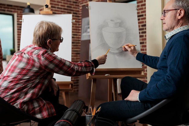 Elderly teacher explaining illustration lesson to student during art course in creativity lesson. Students drawing vase sketching model developing new skill for personal growth in community center