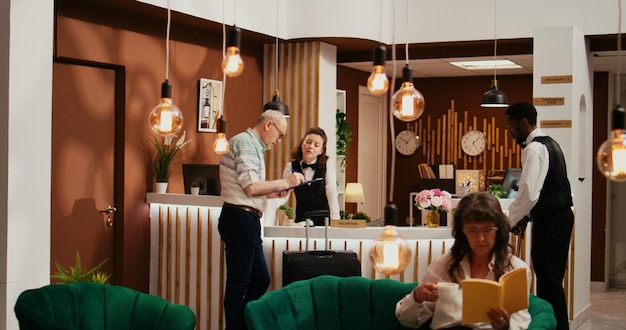 Elderly tourist signing check in papers at front desk, going through registration formalities with hotel staff at holiday retreat. Traveller registering at all inclusive resort, hospitality industry.