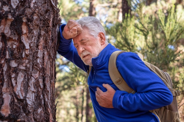 Elderly whitehaired man hiking in the mountains leans against a\
tree trunk to catch his breath placing a hand on his heart senior\
grandfather doesn\'t feel well