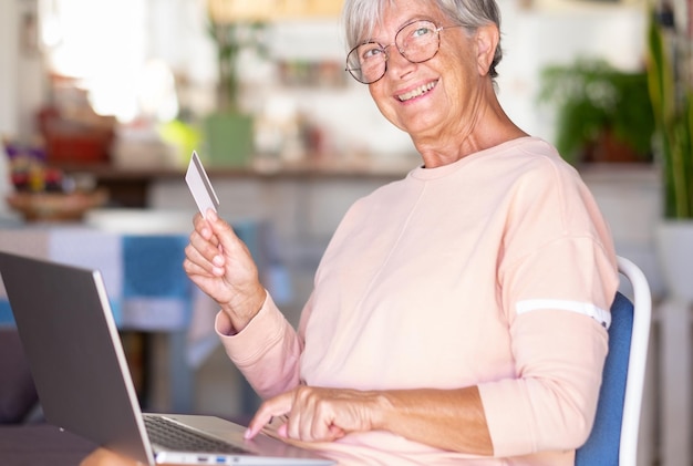 검은 금요일 온라인 쇼핑을 즐기는 노트북으로 탐색하는 노인 여성 전자 상거래 구매를 위해 신용 카드를 사용하여 기분이 좋은 집에서 즐겁고 웃는 노인 여성