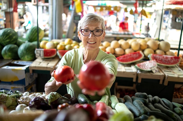 Пожилая женщина покупает свежие органические овощи на рынке для здорового питания.