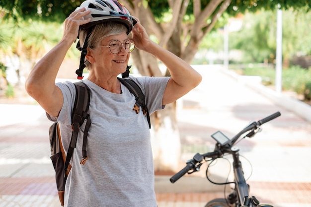 Elderly woman cyclist standing in the urban park next to her\
electric bicycle while taking off her helmet concept of sport\
healthy lifestyle and sustainable mobility