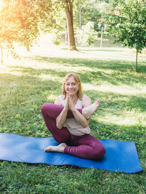 An elderly woman does yoga in the park on a mat. Performs complex Asana exercises, leads a healthy lifestyle. does stretching.