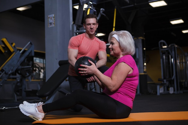 Elderly woman doing medicine ball situps exercising with personal trainer at gym