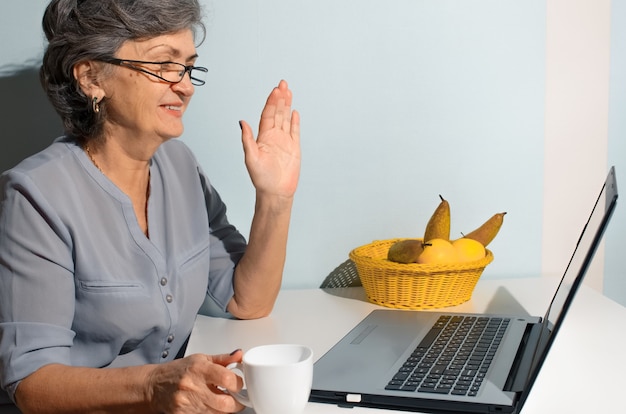 Elderly woman drinking coffee and talking online on laptop at home. Video call concept, new normal, self-isolation