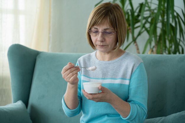 Elderly woman eats yogurt sitting on the couch at home