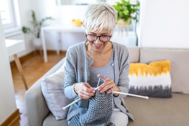 眼鏡をかけた年配の女性が自宅のソファに座って、愛する人のための編み針と糸編みの服、好きな活動と娯楽、引退した静かなのんきな生活の概念を持って笑顔