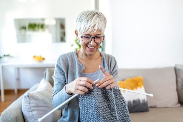 Elderly woman in glasses sit on couch at home smile holding knitting needles and yarn knits clothes for loved ones, favorite activity and pastime, retired tranquil carefree life concept