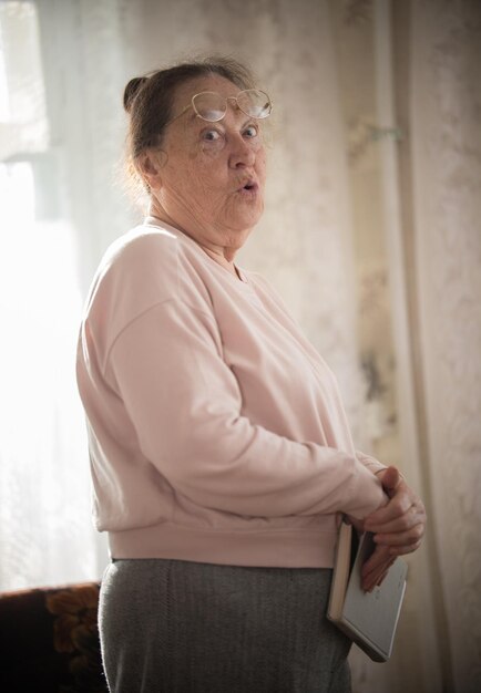 Photo an elderly woman in glasses standin on the background of the window with a book in her arms