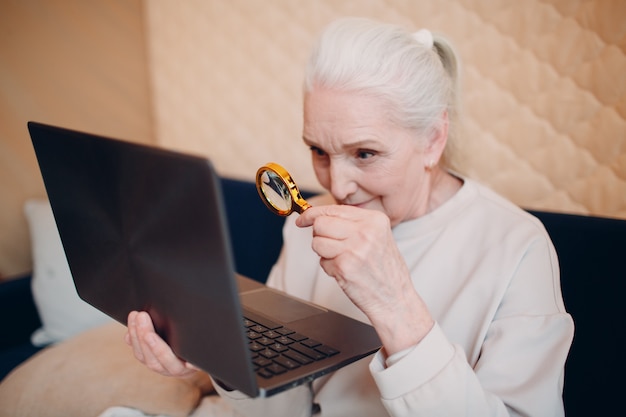 노인 여성 할머니는 돋보기로 노트북 컴퓨터와 인터넷에서 집에서 일하는 법을 배웁니다