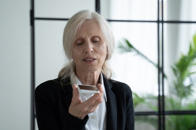 Foto donna anziana che tiene uno smartphone mentre registra un messaggio vocale alla sua amica un moderno impiegato di ufficio anziano tiene uno smartphone alla bocca e registra un messaggio vocale