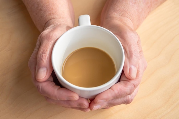 Elderly woman holds morning coffee with milk in hands, on a wooden table.