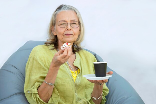 Elderly woman is resting in beanbag chair on veranda of summer\
cafe holding cake and paper cup