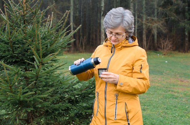 Elderly woman pouring hot drink from thermos outside in forest. Senior woman in yellow raincoat on a walk in cold weather