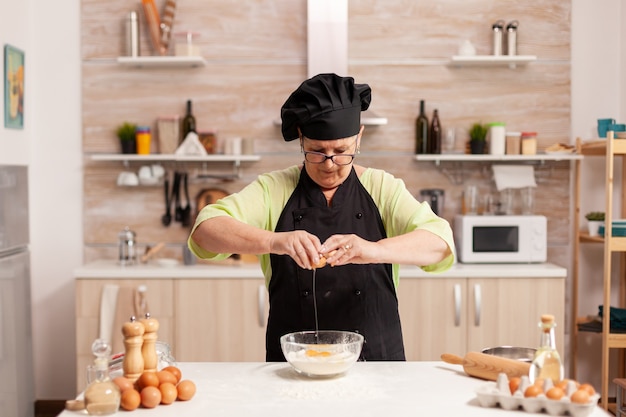 Elderly woman preparing dough cracking eggs over wheat flour following traditional recipe. Elderly pastry chef cracking egg on glass bowl for cake recipe in kitchen, mixing by hand, kneading.