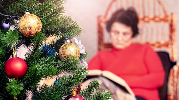 An elderly woman reads a book the Bible in a wicker chair in front of a Christmas tree Celebrating Christmas alone